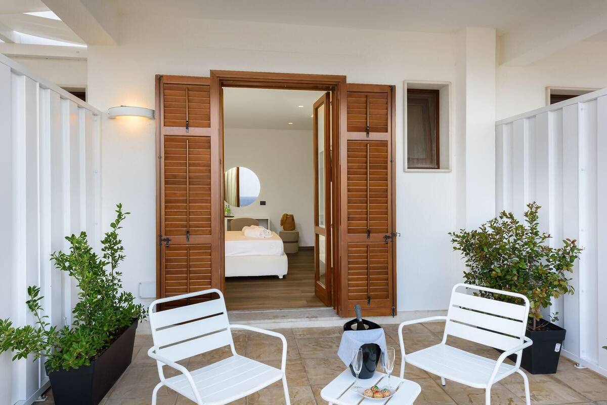 A hotel room with a bed, a mirror, and a view of the sea. There are two white chairs and a table with a bottle and glasses.