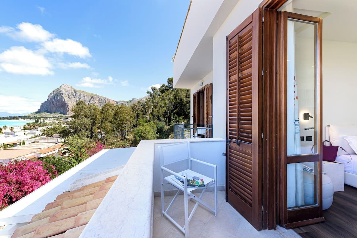 A balcony with a white chair, a small table, and a view of the sea and mountains.