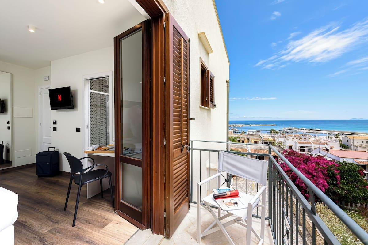 A balcony with a view of the sea, a TV mounted on the wall, and a table with chairs.