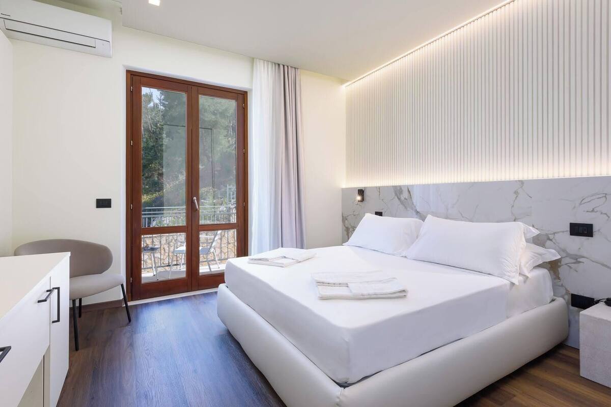 A modern hotel room with a large bed, a desk, and a view through a glass door.
