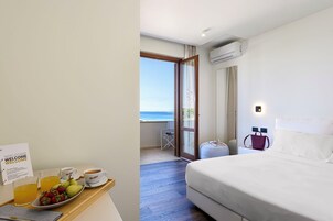Habitación de hotel con una cama, bandeja de desayuno con fruta y café, vista al mar a través de una puerta de vidrio y un equipo de aire acondicionado en la pared.