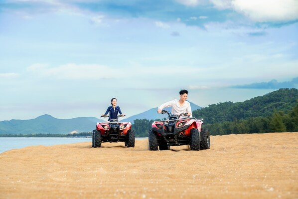 Two people riding ATVs on a sandy beach with a lake and mountains in the background.