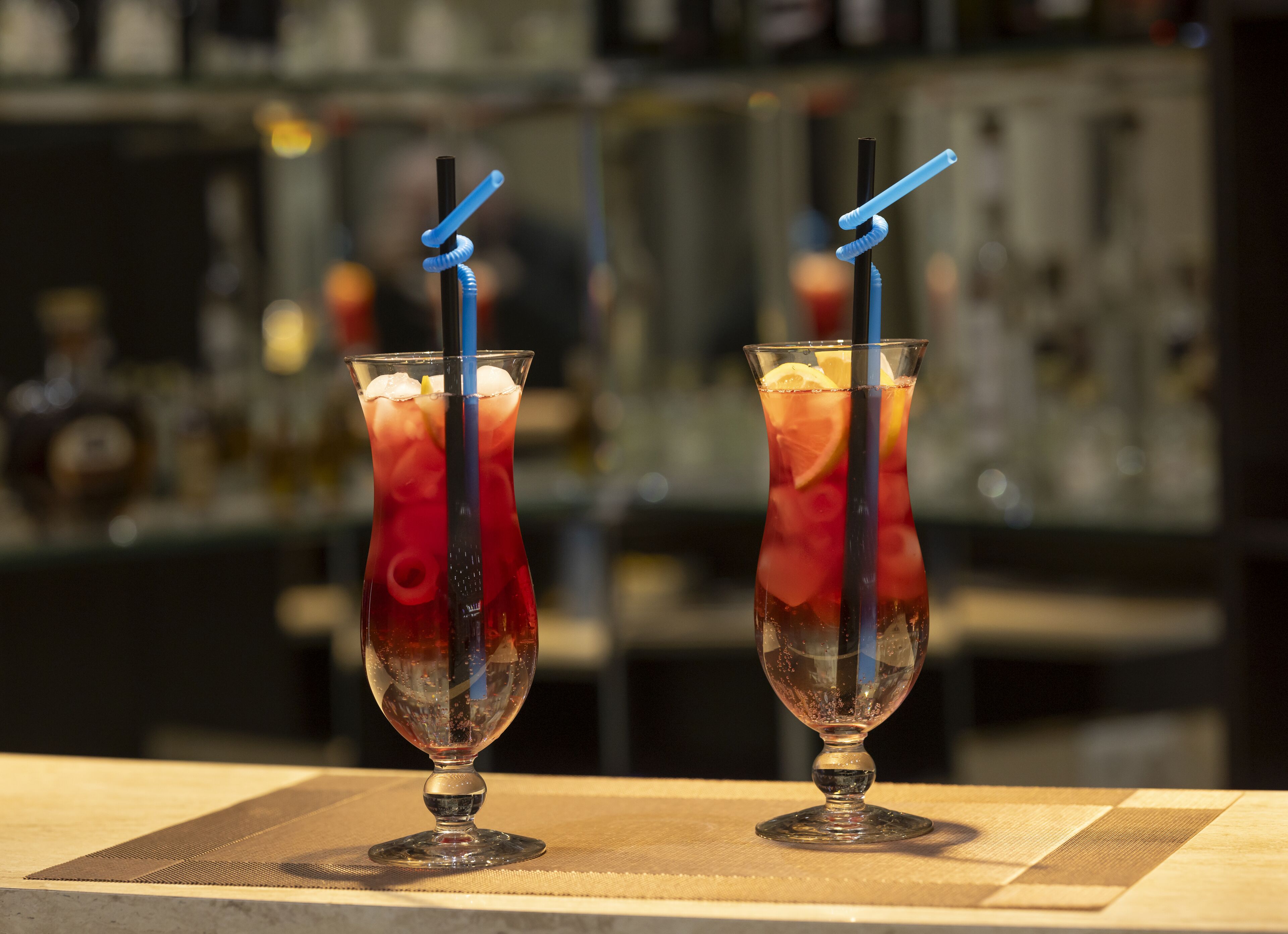 Two tall glasses of red beverage with lemon slices and a blue straw, placed on a table with a patterned tablecloth.