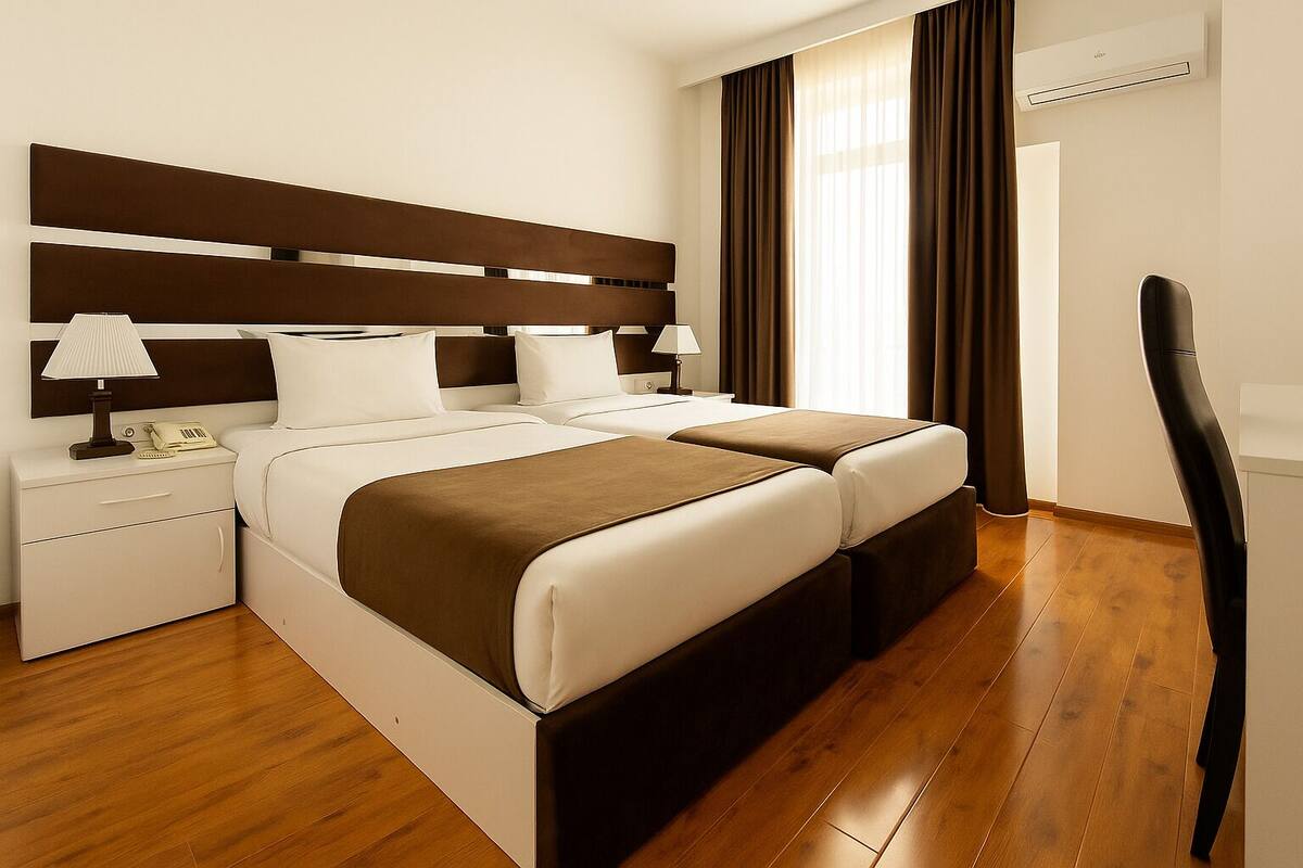 Una habitación de hotel moderna con una cama grande, dos mesitas de noche con lámparas, piso de madera y un ventanal con cortinas.