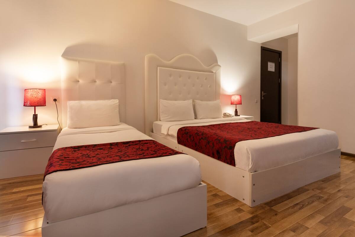 Habitación de hotel con dos camas, cada una con colcha estampada roja, ropa de cama blanca y cabeceros acolchados. Hay mesitas de noche con lámparas, piso de madera y una puerta al fondo.