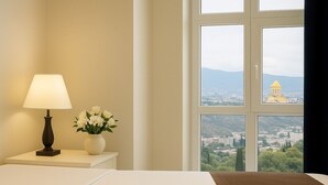 A hotel room with a bed, a nightstand with a lamp and flowers, and a window with a cityscape view. - Hotel Grand Palace Tbilisi (Tbilisi)