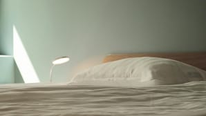 Cama bem arrumada com travesseiro branco e cabeceira de madeira. Uma luminária de cabeceira está à esquerda.