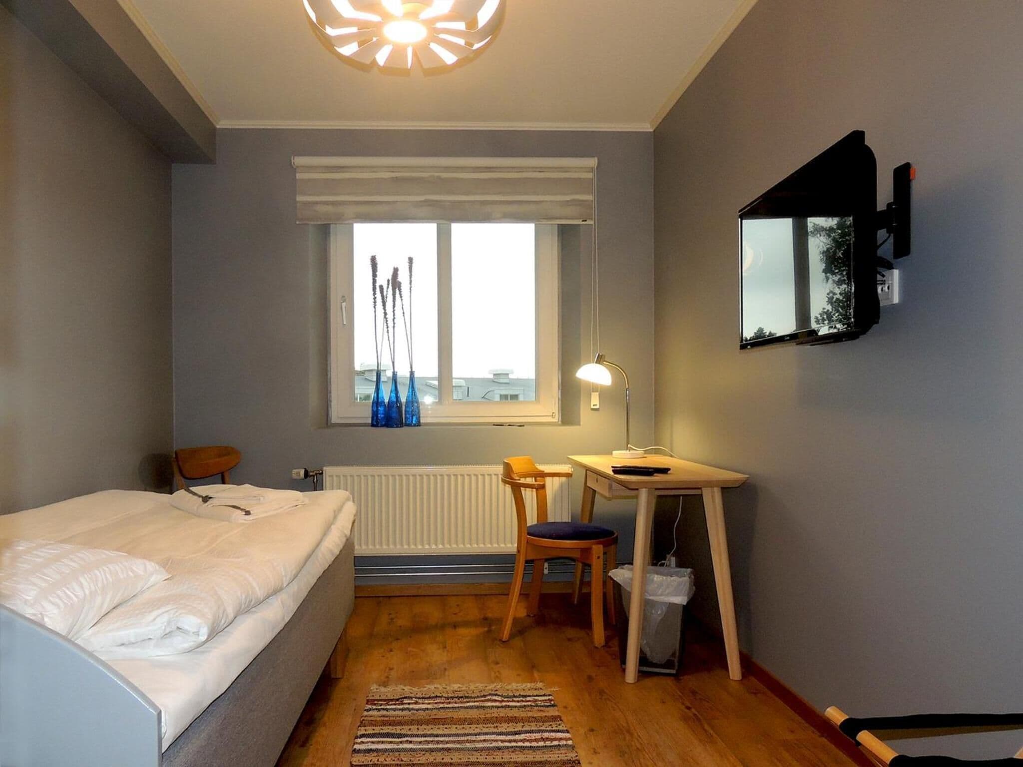 Un dormitorio con cama, escritorio, silla, radiador, ventana y lámpara de techo.