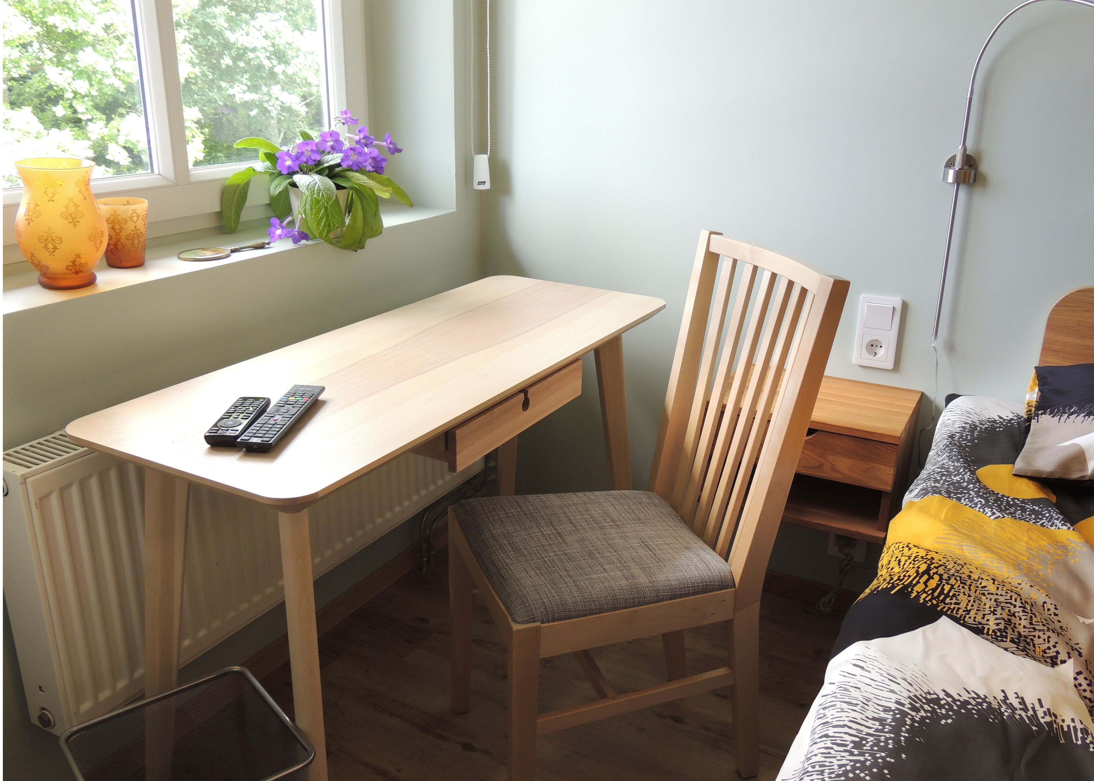 A room with a wooden desk, a chair, a bed with patterned bedding, a radiator, a window with a view of greenery, and a vase with flowers.