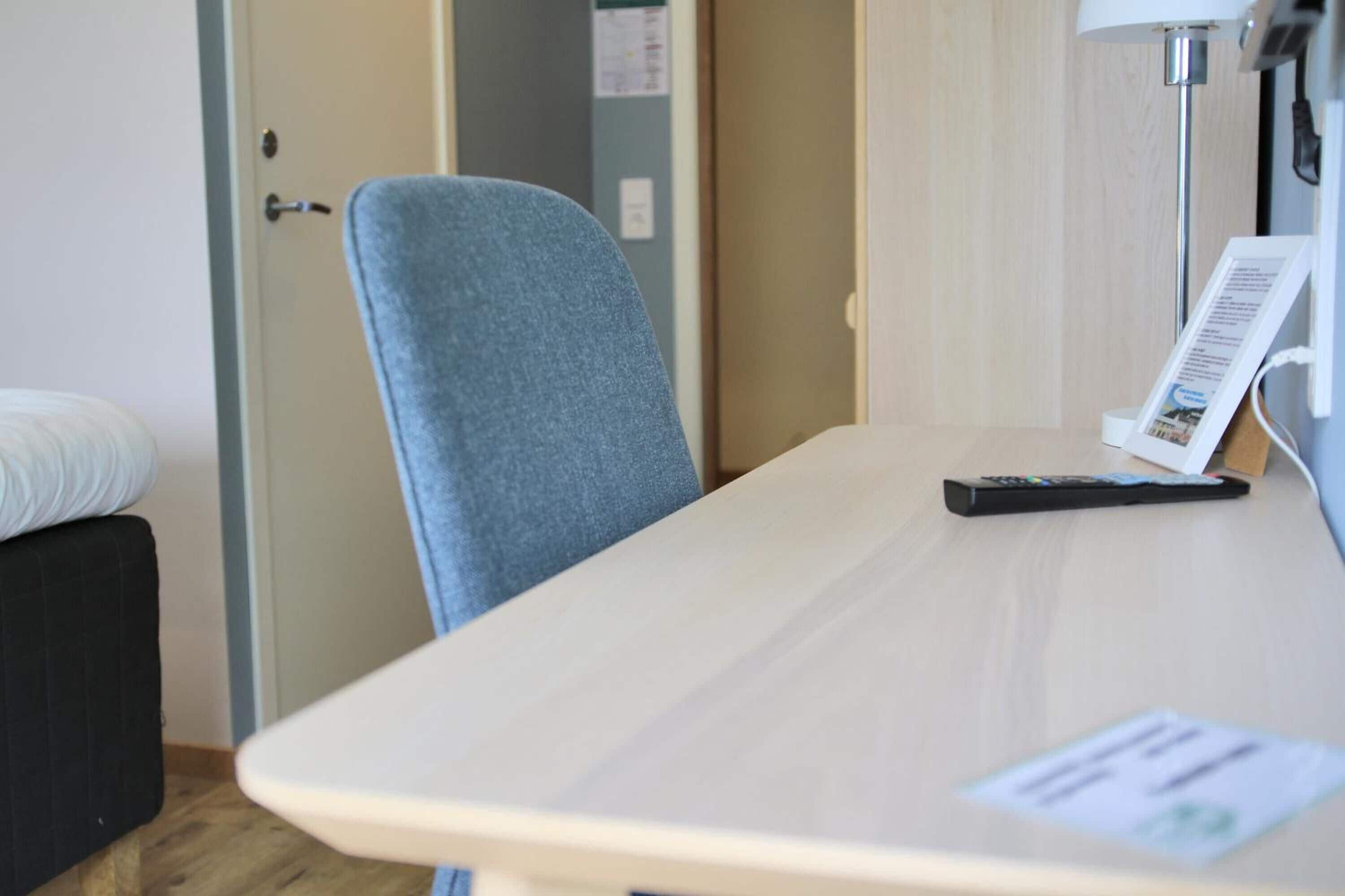 A hotel room with a bed, a desk with a lamp, a chair, and a remote control.