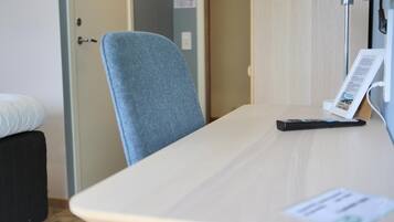 A hotel room with a bed, a desk with a lamp, a chair, and a remote control.