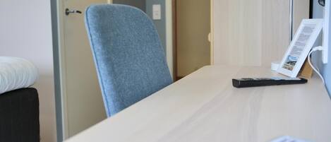 A hotel room with a bed, a desk with a lamp, a chair, and a remote control.