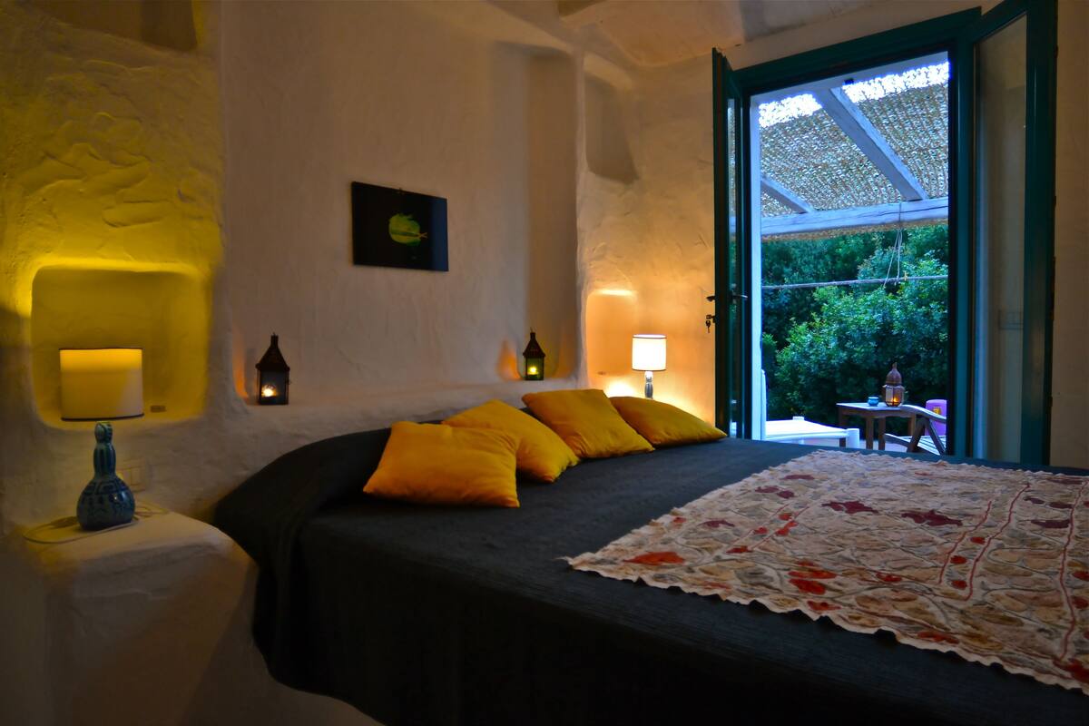 Ein Schlafzimmer mit einem Bett, gelben Kissen, einem Nachttisch mit Lampe und Blick auf Grün hinter einer offenen Tür.