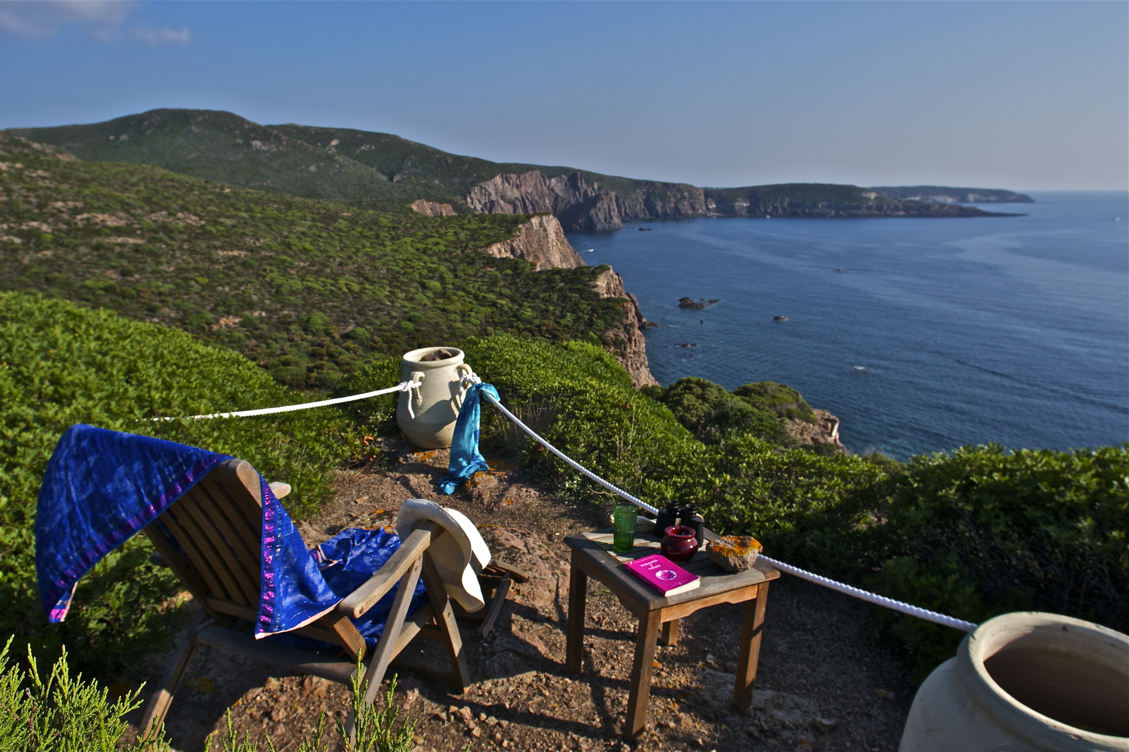 A chair with a blue towel, a wooden table with a book and a cup, and a rope tied to a pot on a rocky seaside view.