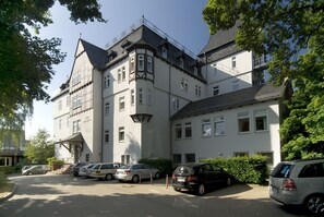 A large, multi-story building with a classic architectural style, featuring a prominent corner tower and multiple windows. There are several cars parked in front, and the building is surrounded by greenery. - Hotel Haus Hainstein (Eisenach)