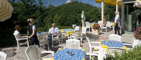 Un patio al aire libre con sillas y mesas blancas, algunas cubiertas con manteles amarillo y azul, y una vista de una colina con un edificio en el fondo.