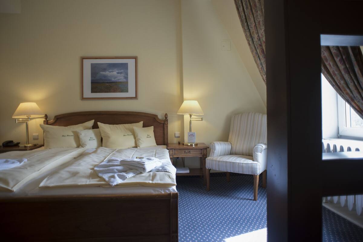 Een hotelkamer met een groot bed, twee bedlampen, een stoel, een nachtkastje en een lijstje aan de muur.