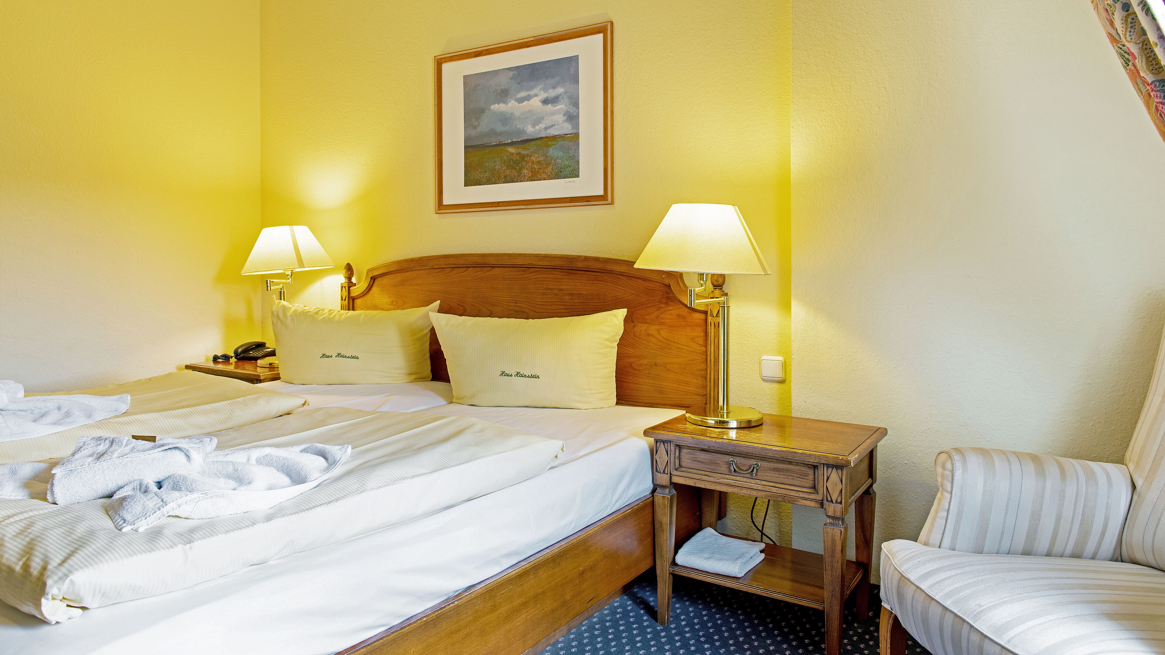 Habitación de hotel con dos camas, cabecero de madera, dos lámparas de noche, un cuadro en la pared y una mesita de noche con lámpara.
