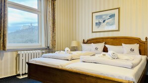 Quarto de hotel com uma cama grande, uma janela com cortinas, um radiador, um quadro emoldurado na parede e uma luminária na mesa de cabeceira.