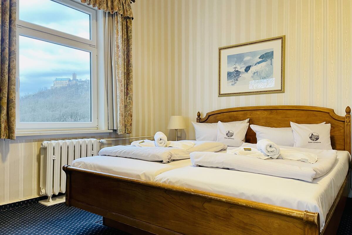 Een hotelkamer met een groot bed, een raam met gordijnen, een radiator, een ingelijste foto aan de muur en een lamp op het nachtkastje.
