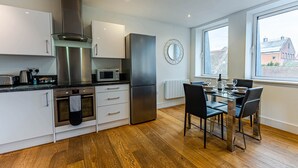 A modern kitchen with stainless steel appliances, a dining area with a table and chairs, and a large window providing a view of buildings outside. - Celador Apartments - Sussex House Serviced Apartments (Reading)