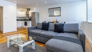 A modern living room with a grey sectional sofa, a glass coffee table, and a kitchen in the background. - Celador Apartments - Sussex House Serviced Apartments (Reading)