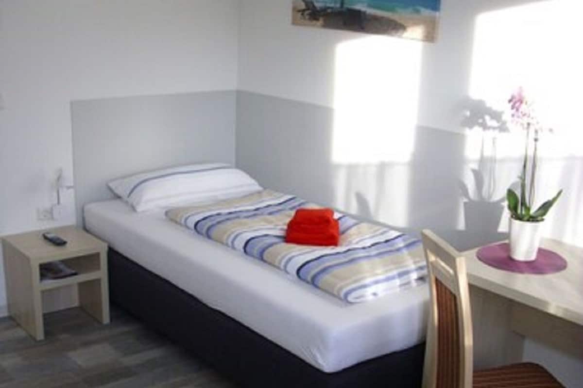 A bedroom with a bed, a nightstand, a desk with a chair, a potted plant, and a painting on the wall.