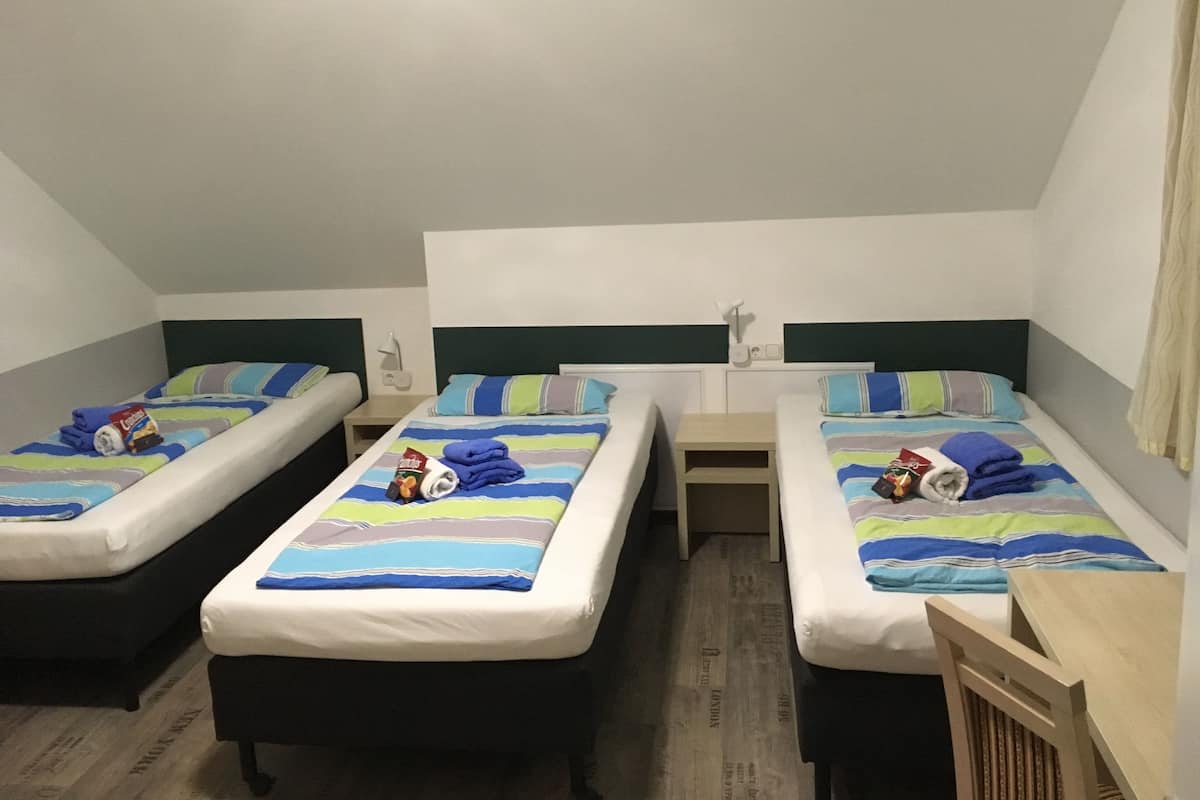 A dormitory room with three single beds, each with a striped blanket and pillows. There are bedside tables with lamps and a small shelf.
