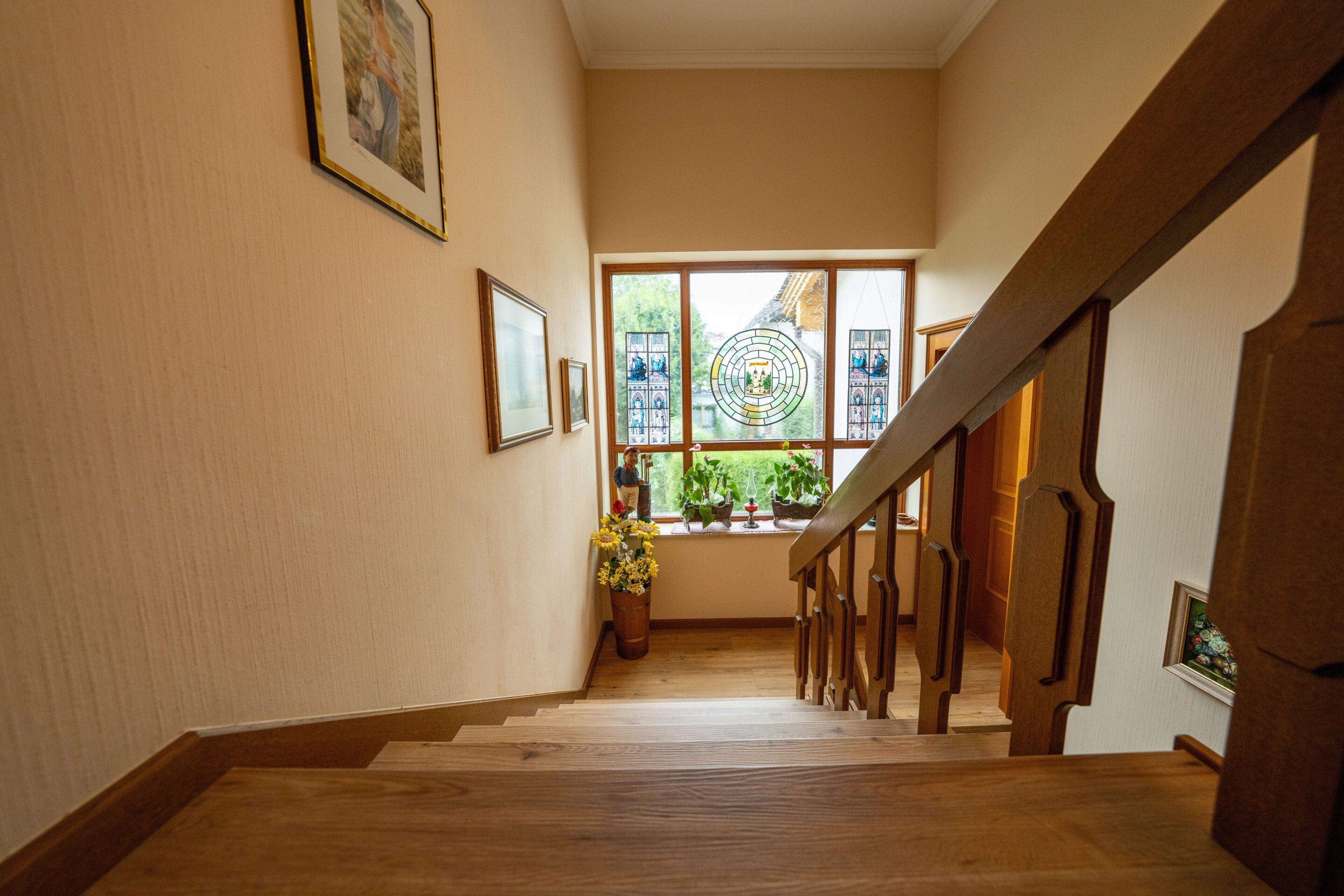 A wooden staircase with a handrail leading to a landing. There are framed pictures on the walls and a window with a view of greenery outside.