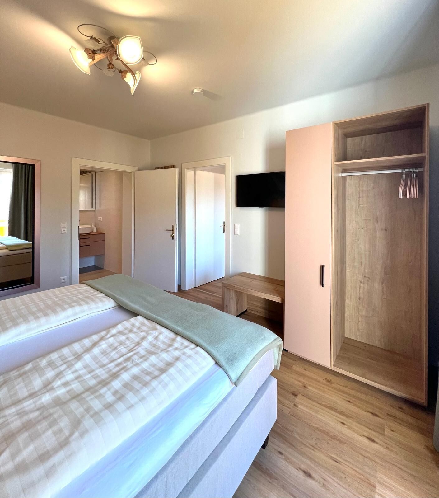 A bedroom with two beds, a wooden wardrobe, a television mounted on the wall, and a ceiling light fixture.
