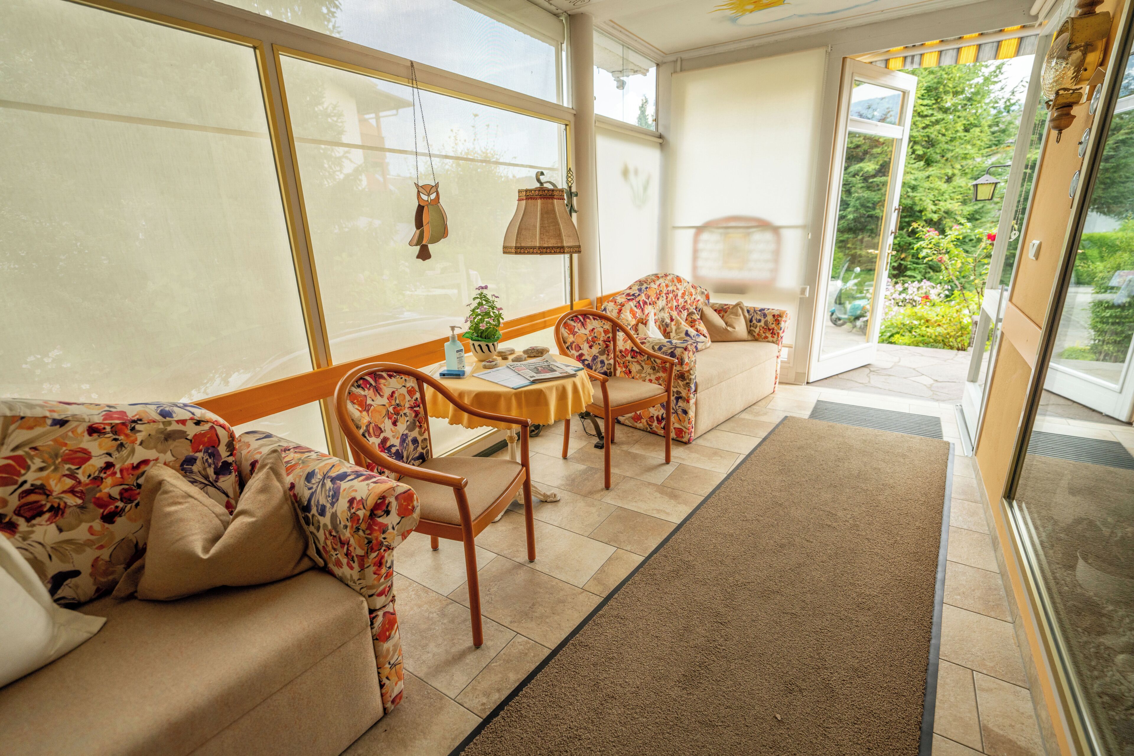 A sunlit room with a sofa, chairs, a small table, and a view of a garden through a glass door.