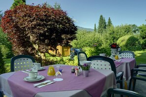 Un área de comedor al aire libre con mesas dispuestas para una comida, rodeada de abundante follaje y un gran árbol con hojas rojas.