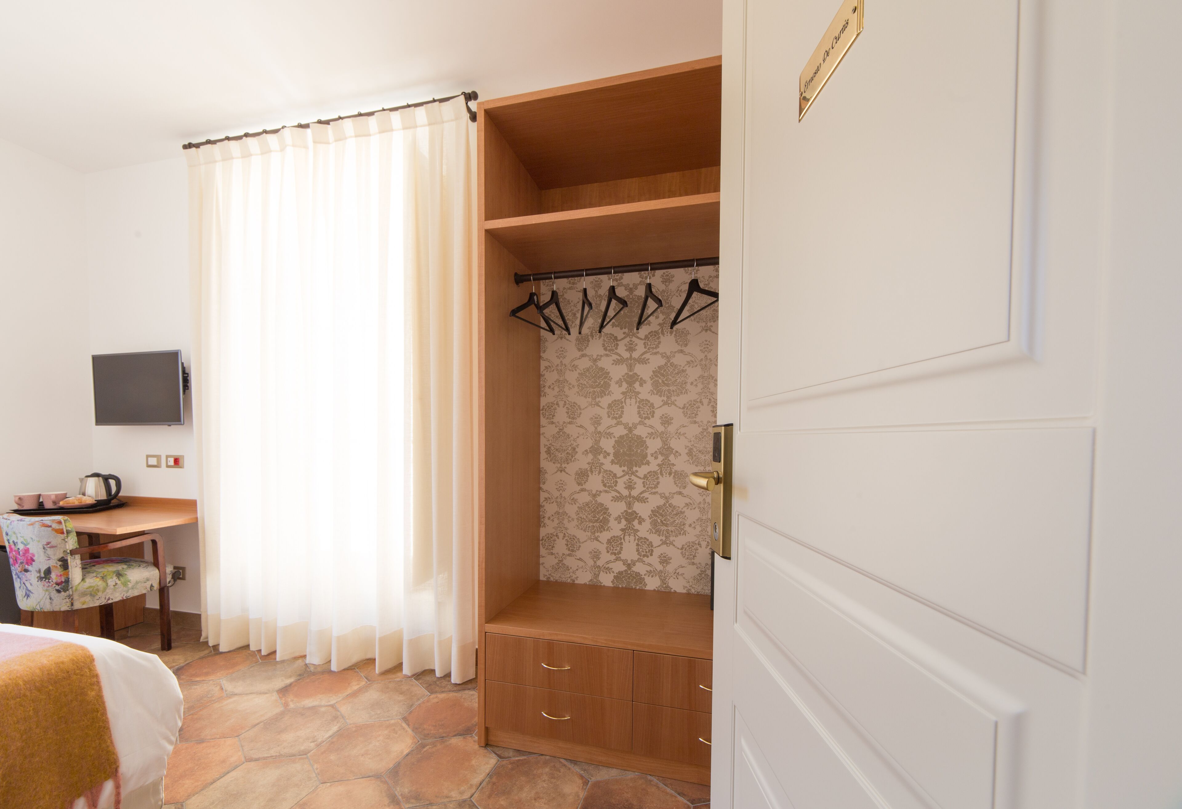 Quarto de hotel com piso de pedra, um banco de madeira, televisão, escrivaninha com chaleira e guarda-roupa com cabides.