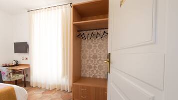 A hotel room with a stone floor, a wooden bench, a television, a desk with a kettle, and a wardrobe with hangers.