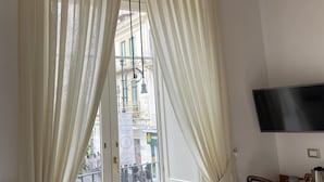 A room with a white door, a chandelier, a table with a TV, and a view of buildings outside. - Residenza Il Campanile (Sorrento)