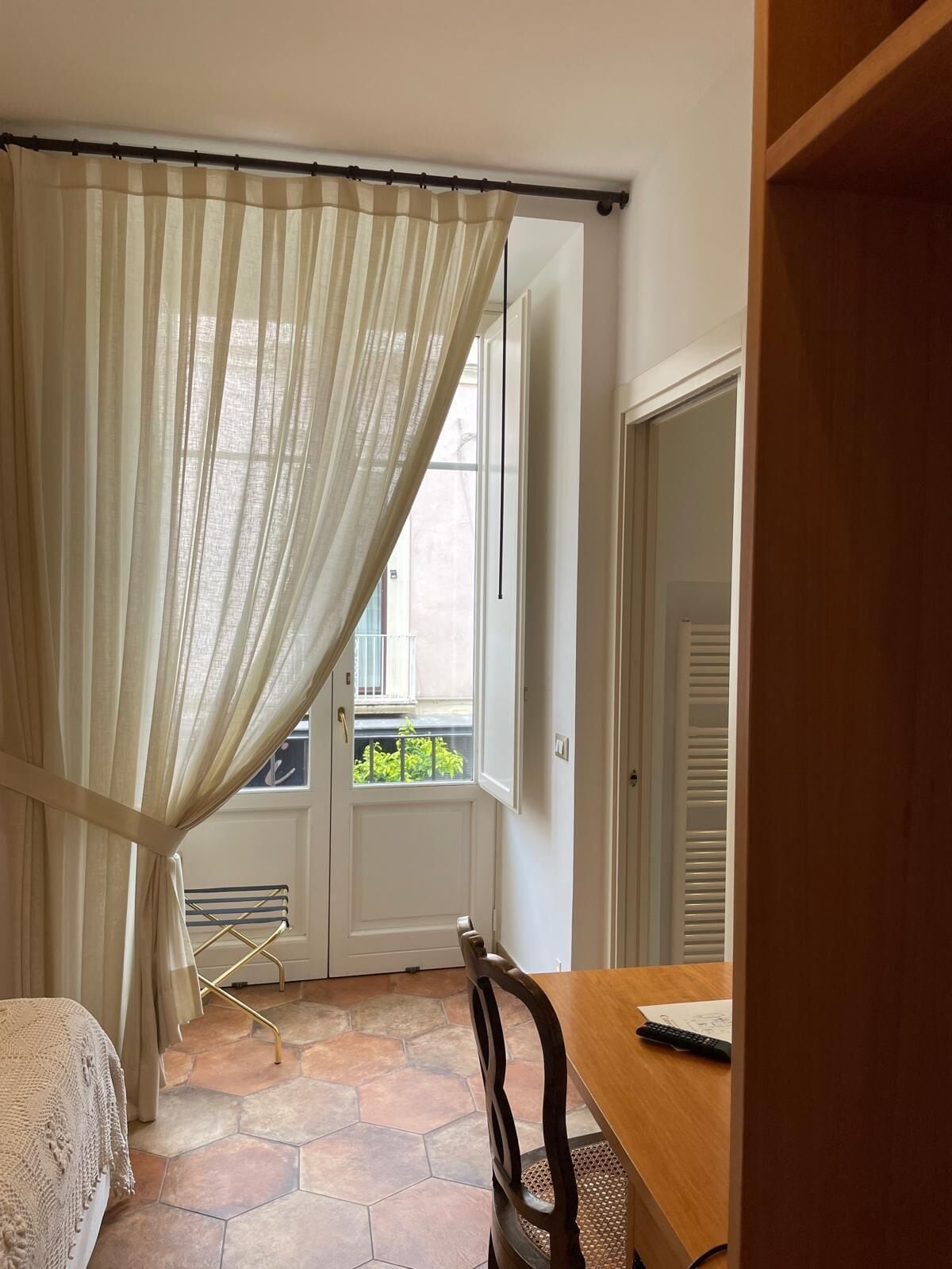 A room with a bed, a wooden table, a chair, a window with a view of greenery, and a door.