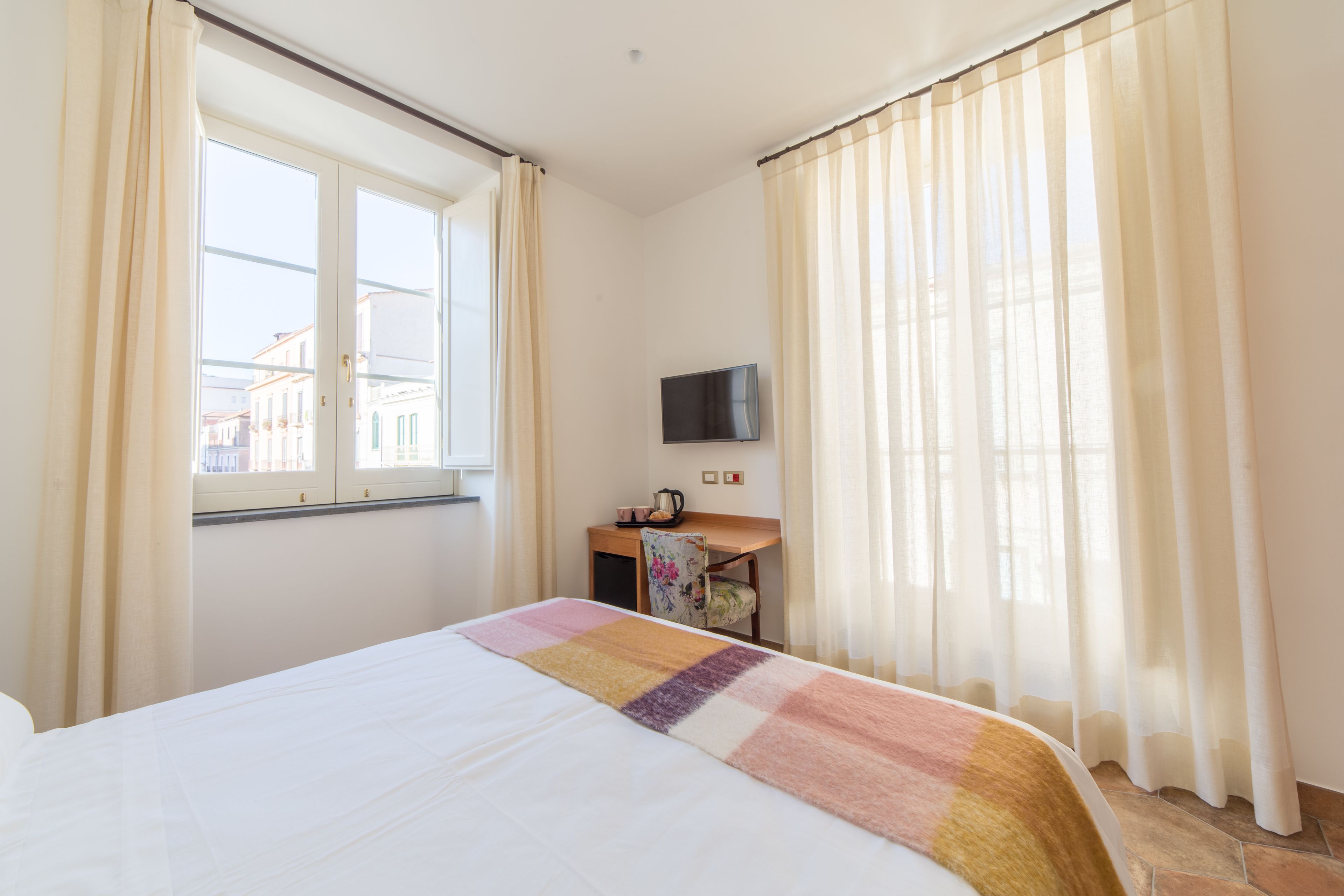 A hotel room with a bed, a desk with a chair, a television, and a window with curtains.