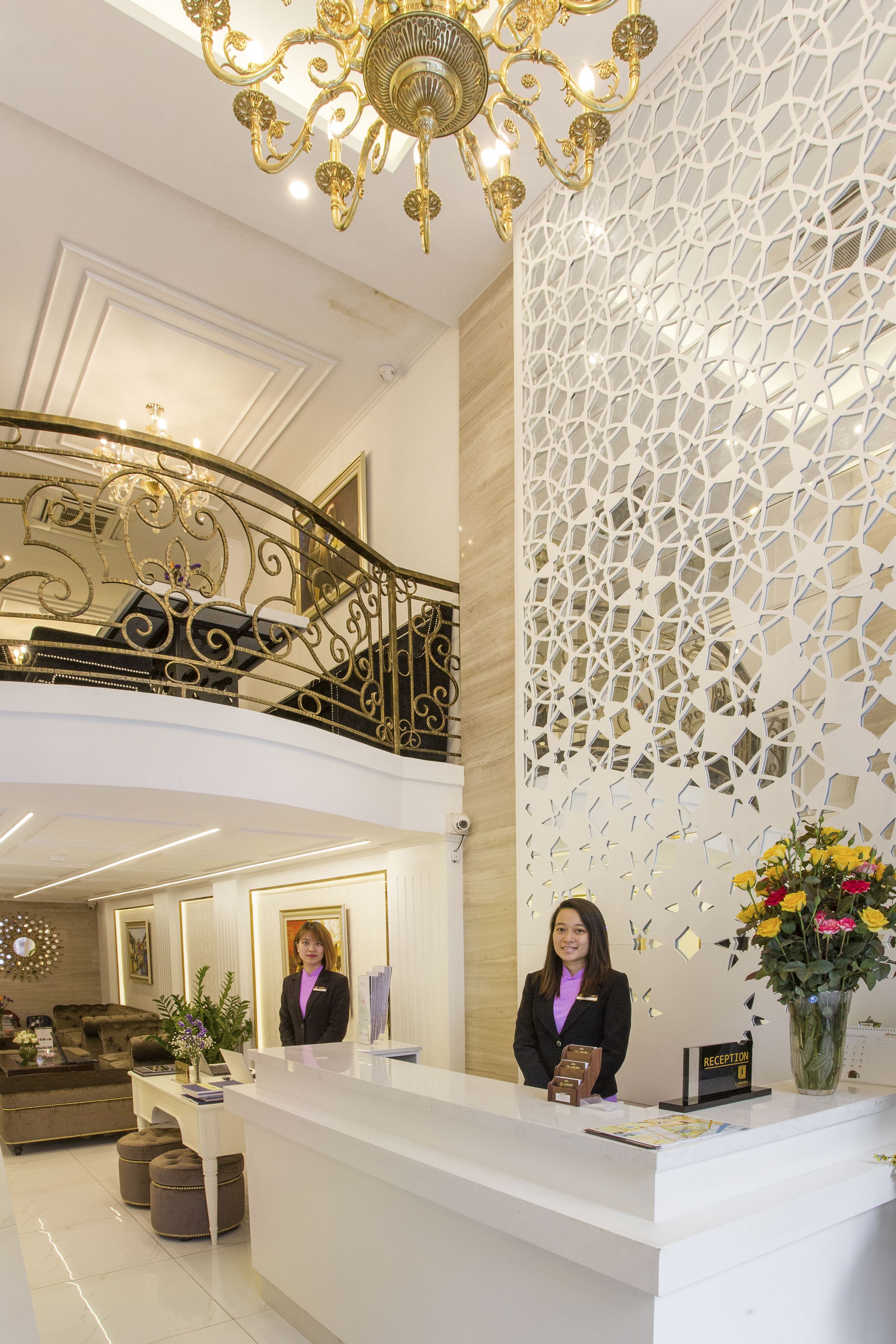 Un hall d’hôtel avec un comptoir de réception blanc, un réceptionniste, un lustre, un escalier et un mur décoratif.