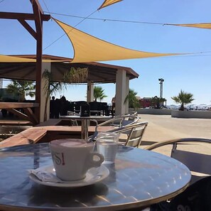 Una mesa de vidrio con una taza y un plato, asientos al aire libre con sillas de metal, y un área techada con mesas y sombrillas.