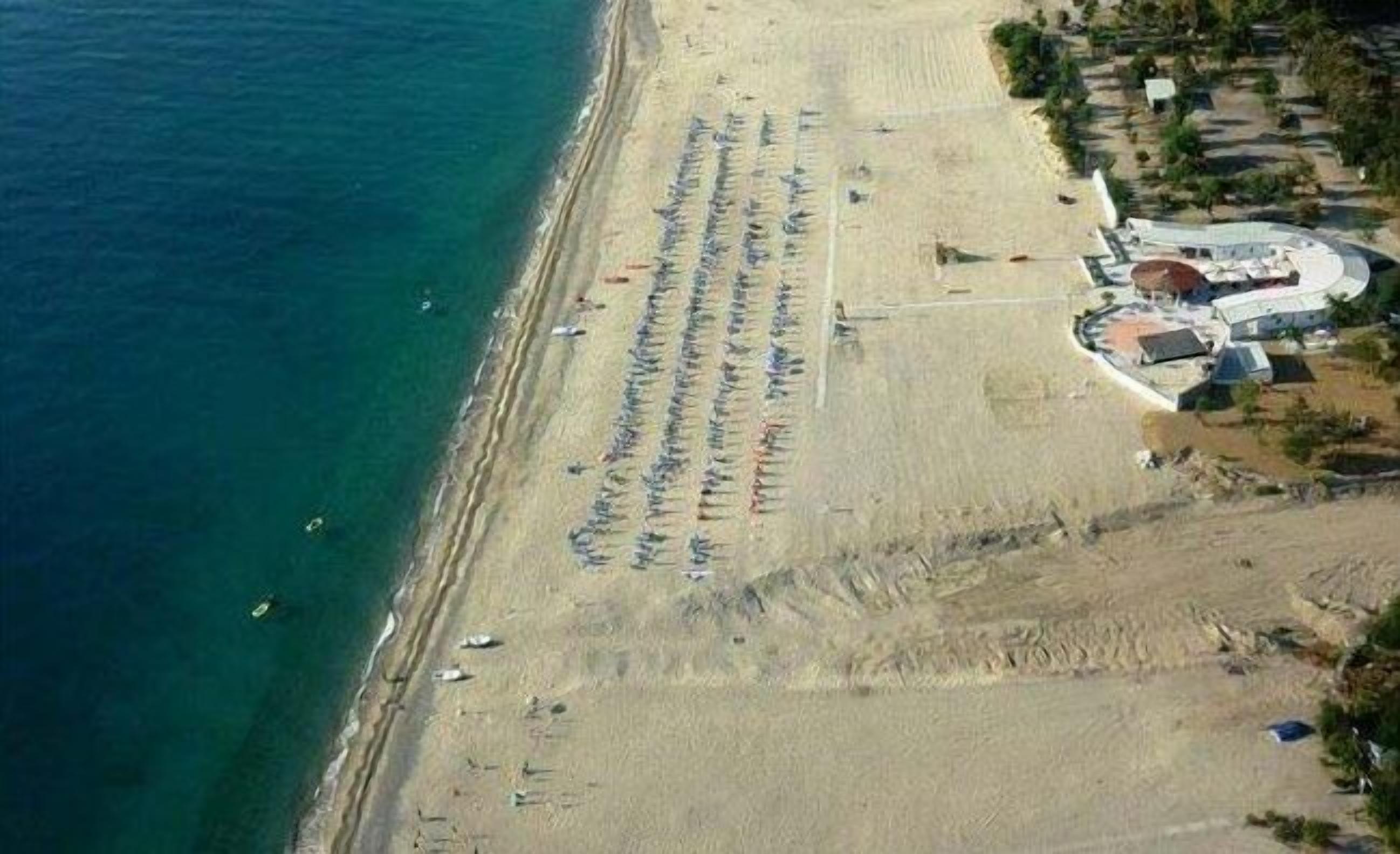 An aerial view of a beach with rows of umbrellas and sun loungers, a few boats, and a building with a curved roof near the water's edge.