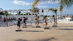 Eine Gruppe von Menschen trainiert auf Podesten in einem Außenbereich mit einer Pergola und Palmen.