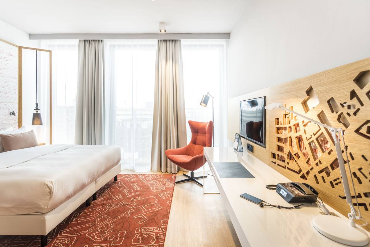 A modern hotel room with a large bed, a red chair, a desk with a phone, and a cityscape view through the window.