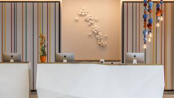 Un comptoir d’accueil moderne, équipé de deux ordinateurs Apple, d’une plante en pot et d’un mur orné de rayures verticales, ainsi que d’un luminaire au design géométrique.