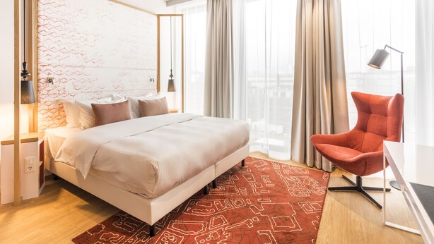 A modern hotel room with a large bed, a red chair, and a desk with a lamp. - Capri by Fraser Berlin (Berlin)