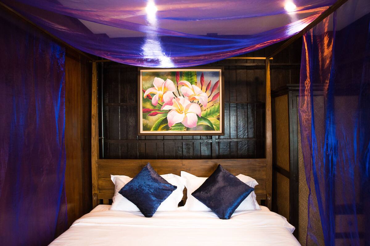 A bed with white linens and two dark blue pillows, a wooden headboard, and a painting of pink flowers on the wall.