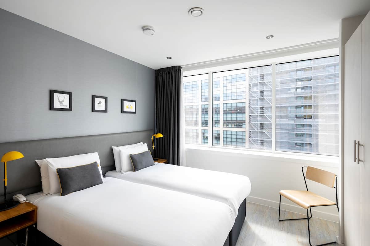 A modern hotel room with a large bed, bedside tables, a chair, and a view of the city through large windows.