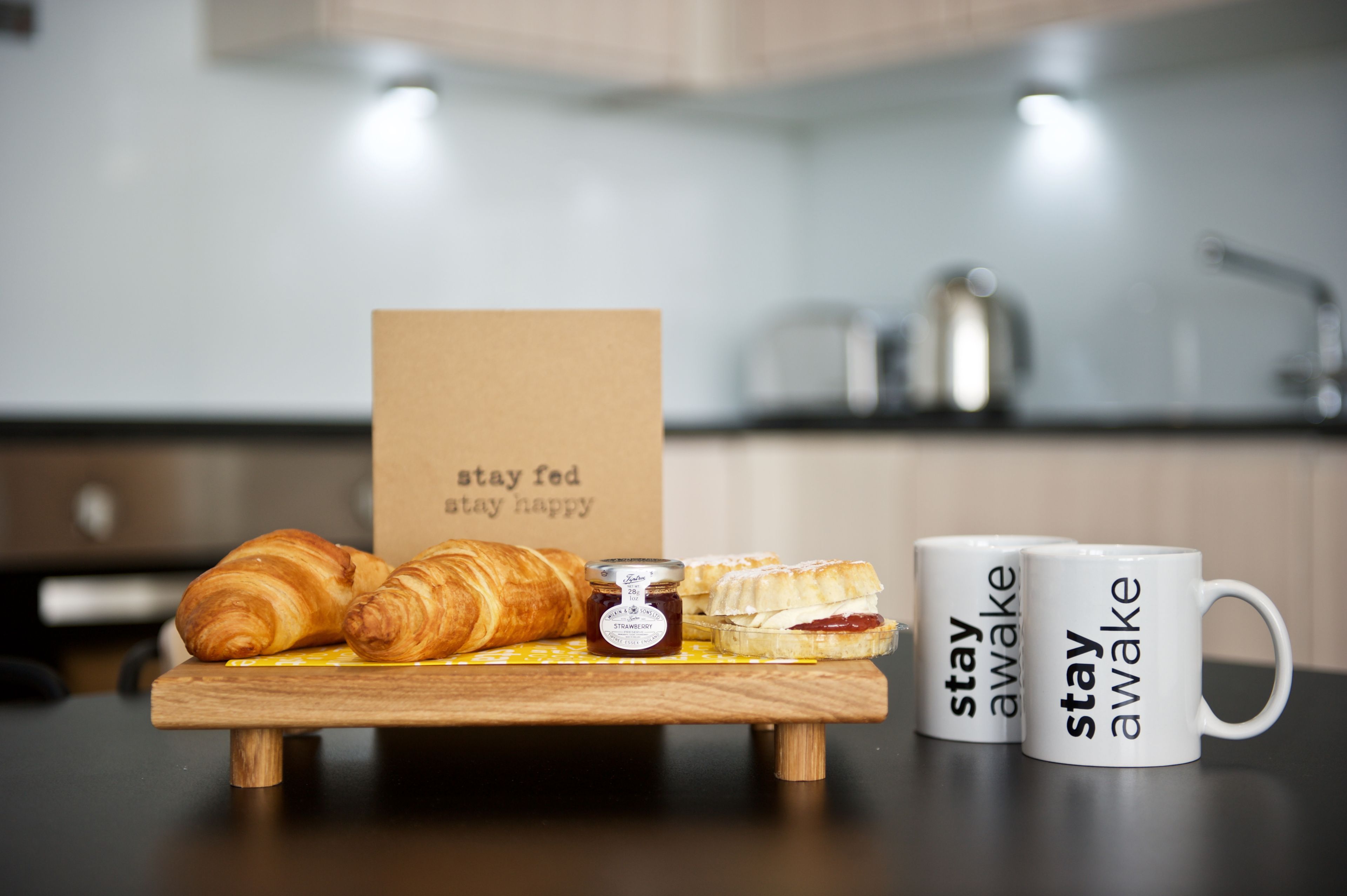A wooden board with croissants, a jar of jam, and a card with the text 'stay fed stay happy' in a kitchen setting.