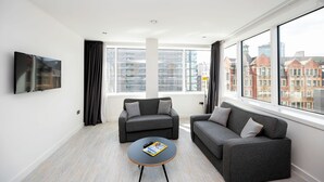 Uma sala de estar moderna com dois sofás cinzentos, uma mesa de centro redonda e uma TV de tela plana na parede. Janelas amplas oferecem vista para prédios da cidade.