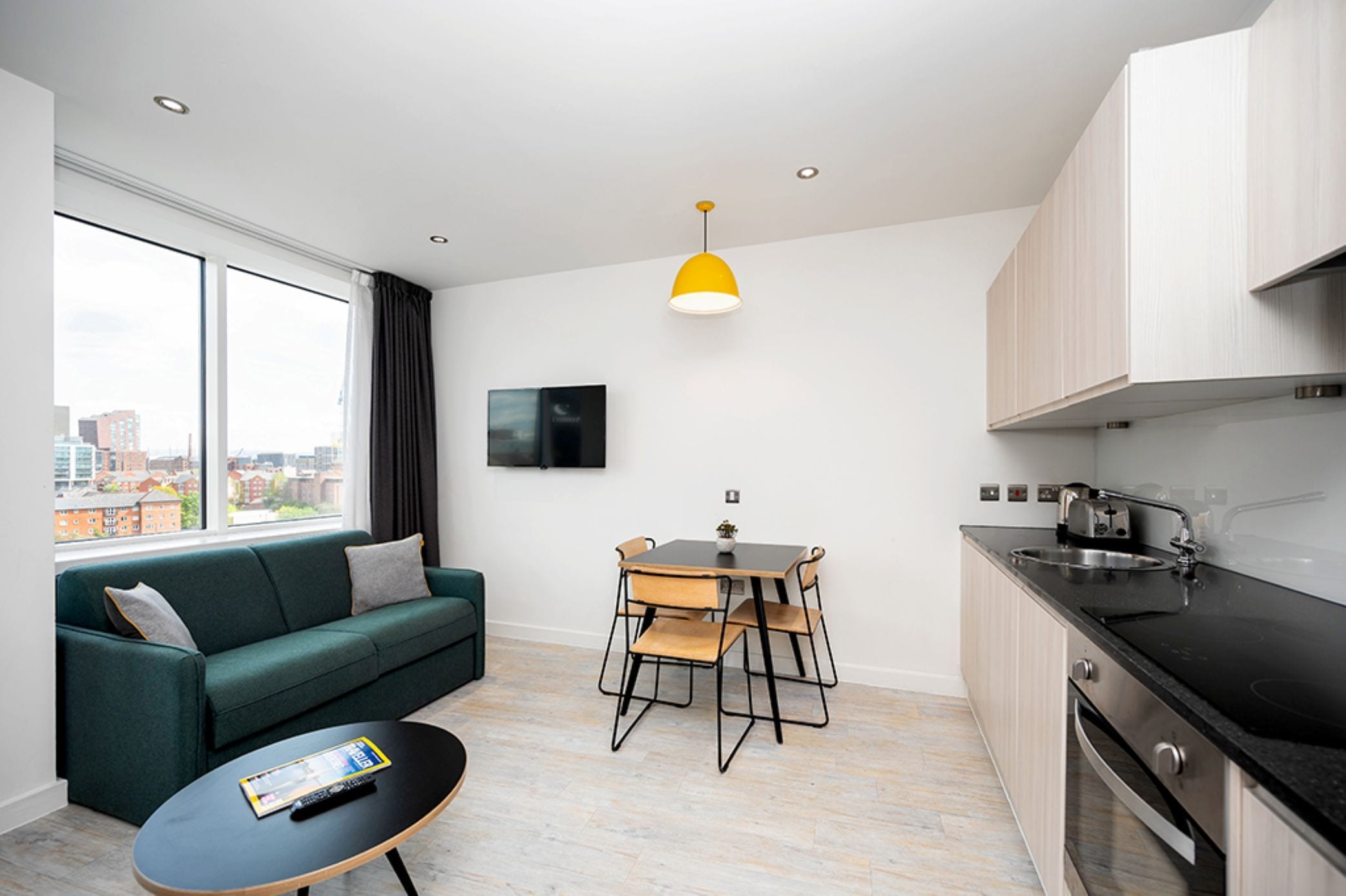 Un salon moderne avec un canapé, une table basse et un coin repas avec des chaises. Il y a une cuisine équipée d’un évier et de placards. Un téléviseur est fixé au mur, et une fenêtre offre une vue sur la ville.