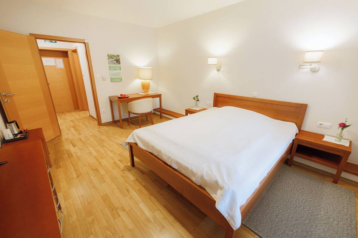 Une chambre d’hôtel avec un lit en bois, un bureau avec une chaise, une petite table avec une lampe et une table de chevet ornée d’un vase de fleurs.